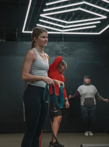 Woman holding resistance bands, others exercising in gym with geometric lighting.