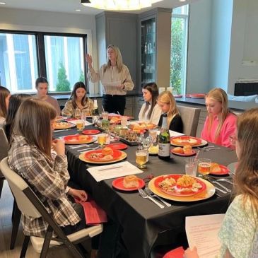 Group of people gathered around a dining table, listening to a standing woman.