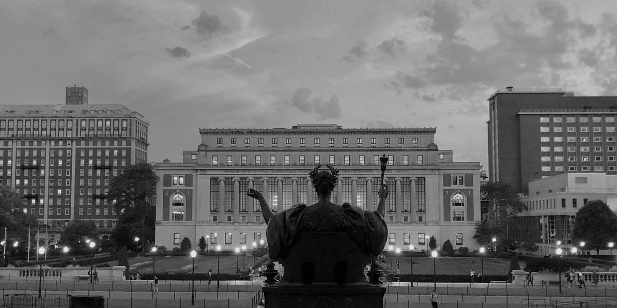 Black and white photo of a statue facing a grand building with lit windows and people walking nearby.