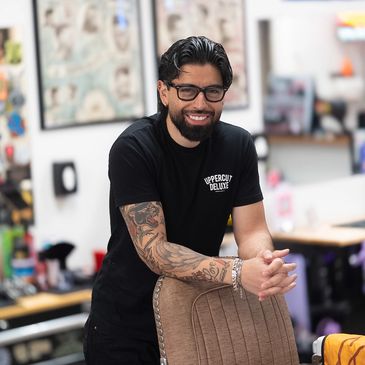 Smiling man with tattoos and glasses leaning on a chair in a casual setting.
