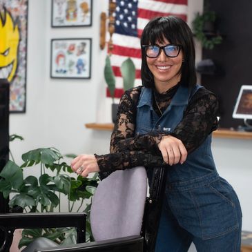 Smiling woman in glasses leaning on a chair with an American flag in the background.