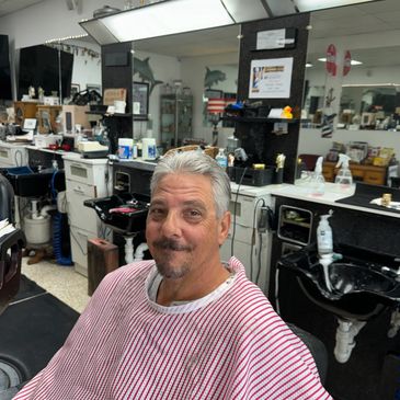 Man with gray hair and goatee in a barber shop chair wearing a striped cape.
