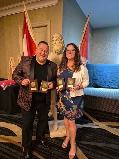 Two people holding awards, standing next to a statue with flags behind them.