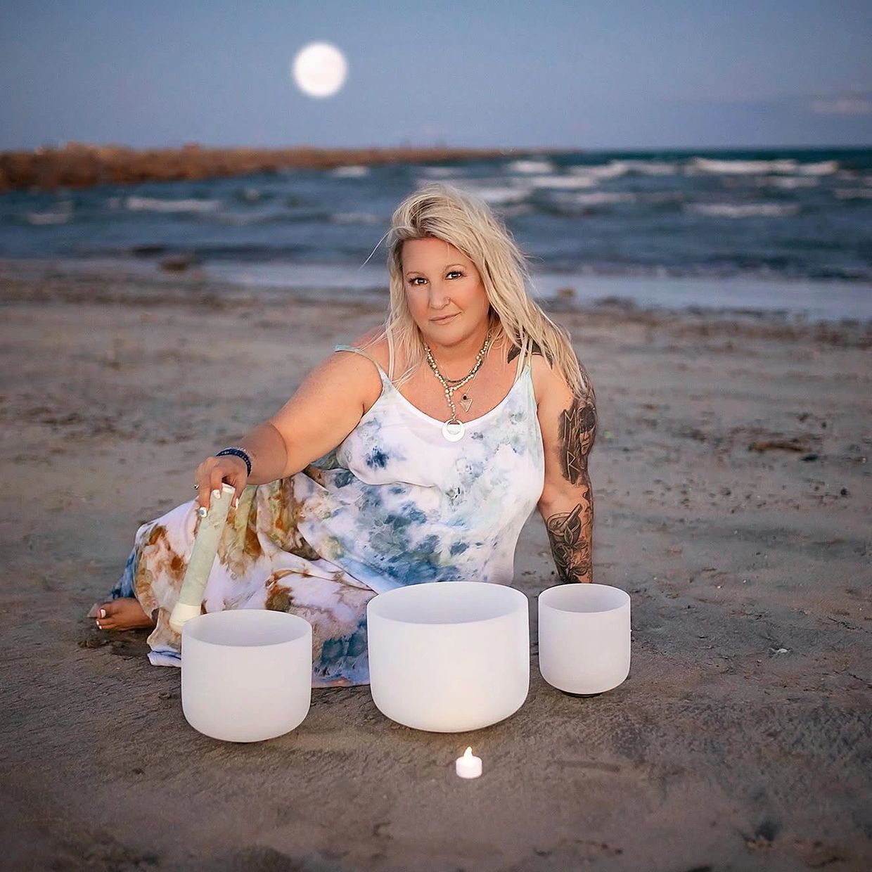 Woman playing crystal singing bowls on the beach at dusk with a full moon.