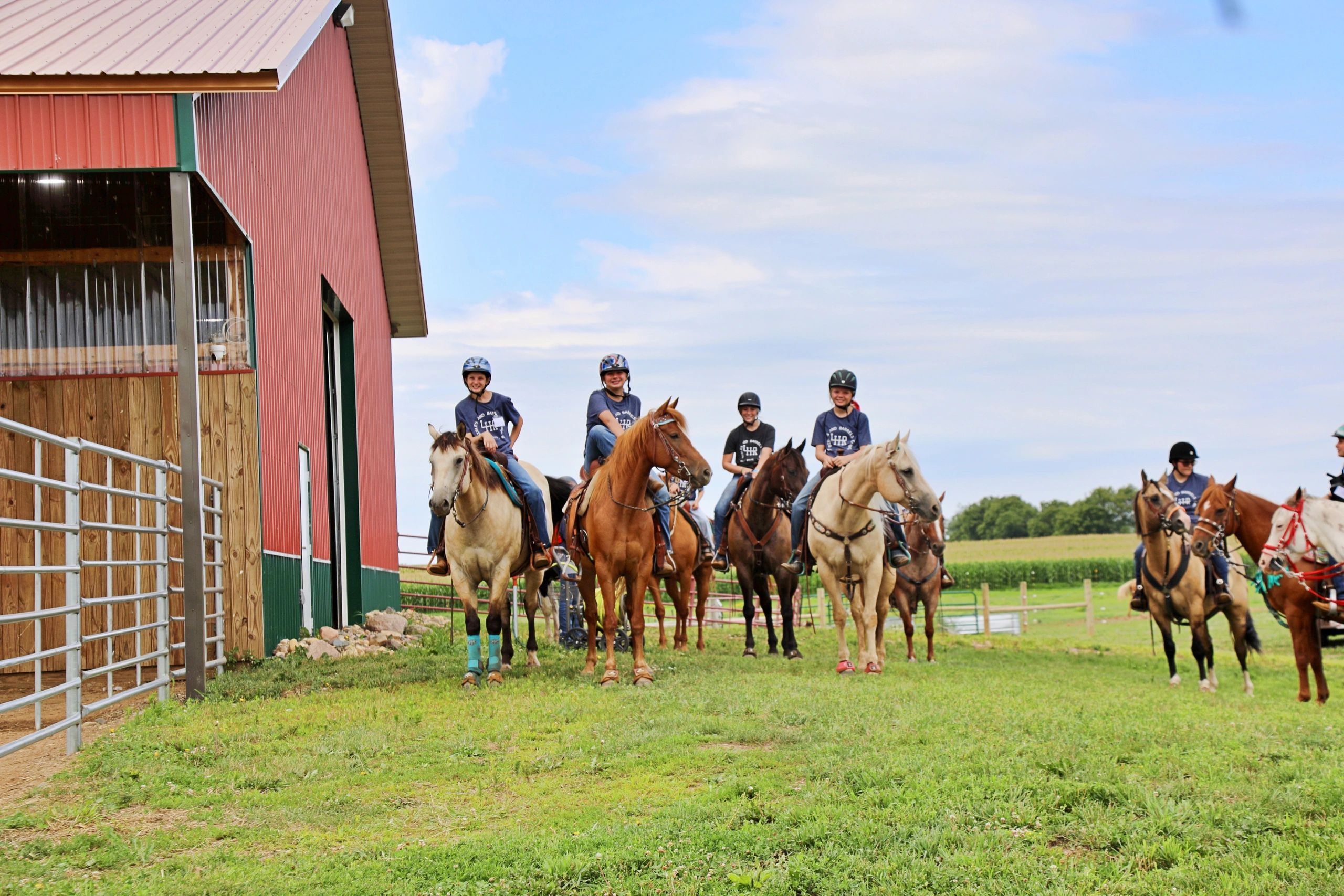 Group of children riding horses near a red barn on a sunny day.
