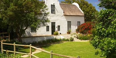 Charming white cottage with a thatched roof surrounded by lush greenery and a wooden fence.