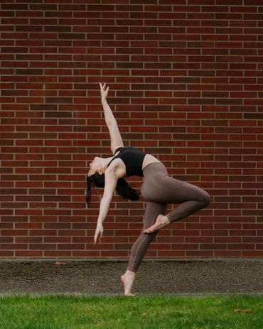 A woman performing a graceful yoga pose outdoors against a brick wall.