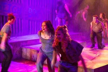 Actors perform energetically on stage under vibrant purple lighting.