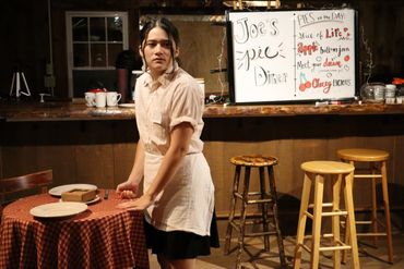 A woman in a diner setting arranging plates on a table with a pie menu behind her.