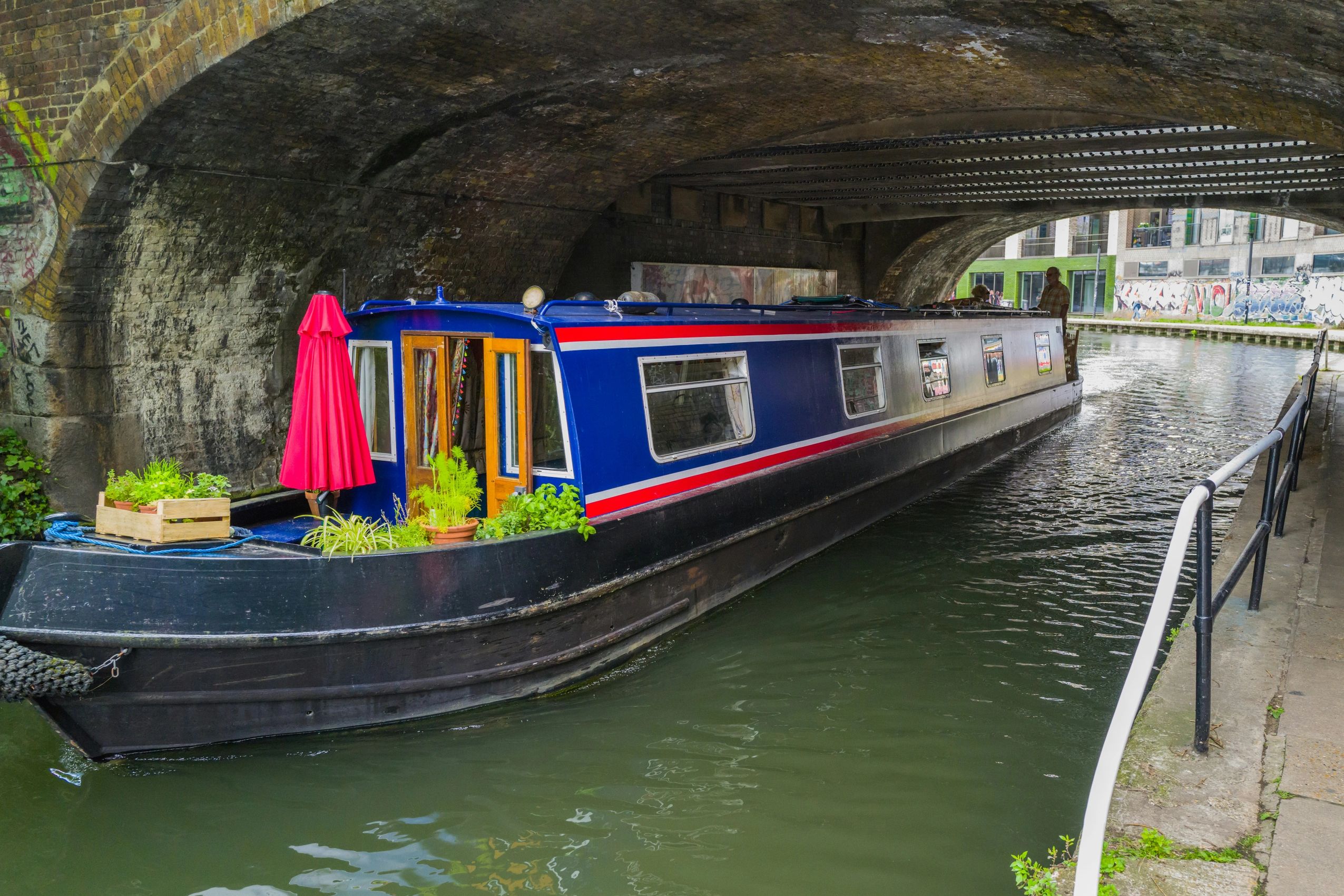 Red and navy coloured canal boat moving on green water, underneath a bridge.