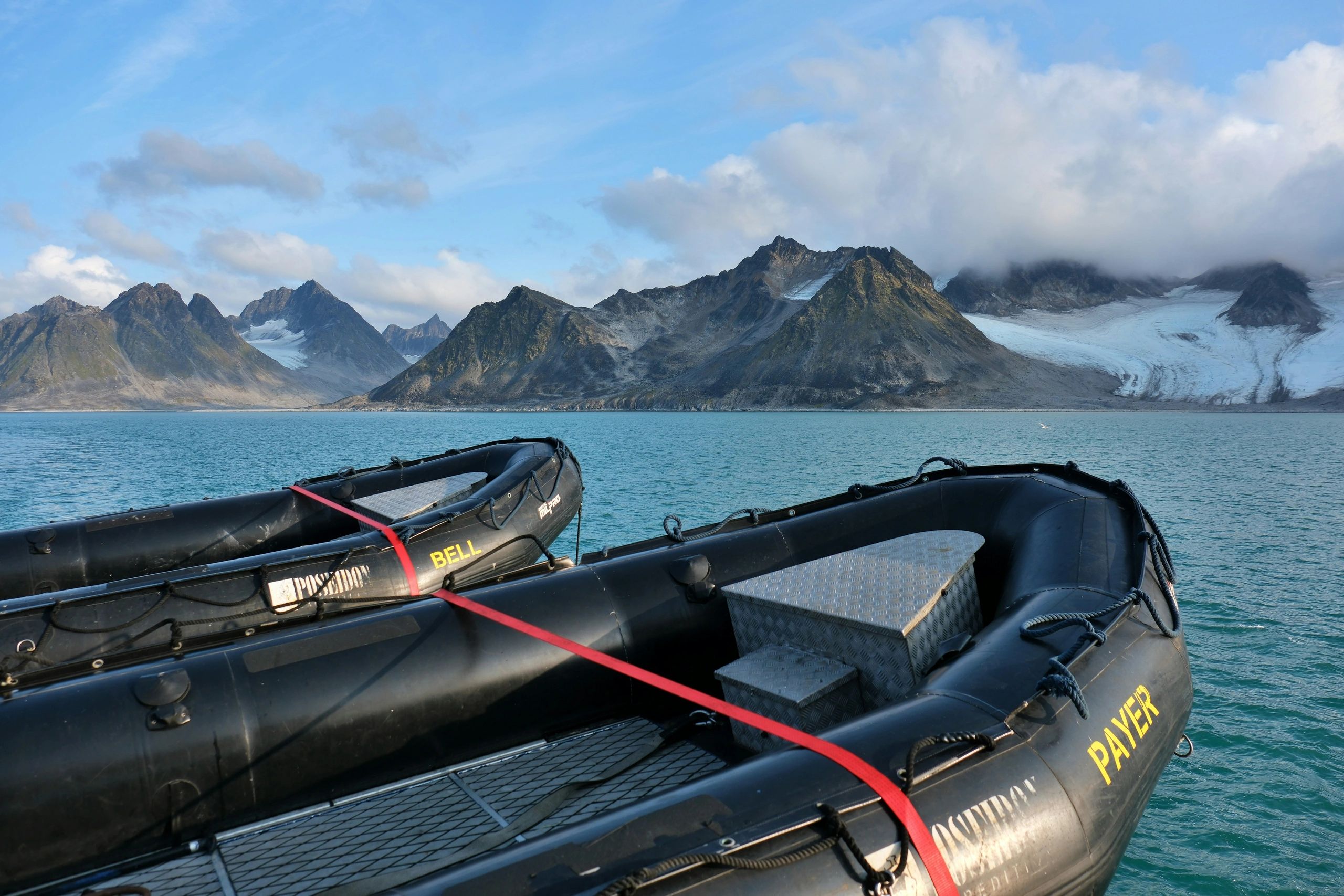 The nose of two black rib boats with a red strap across the bow, and huge, glacier filled mountains 