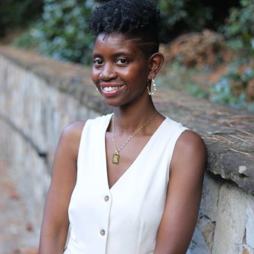 Smiling woman in white sleeveless top by a stone wall.