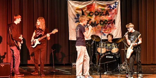 A band performs onstage with instruments and a colorful banner.