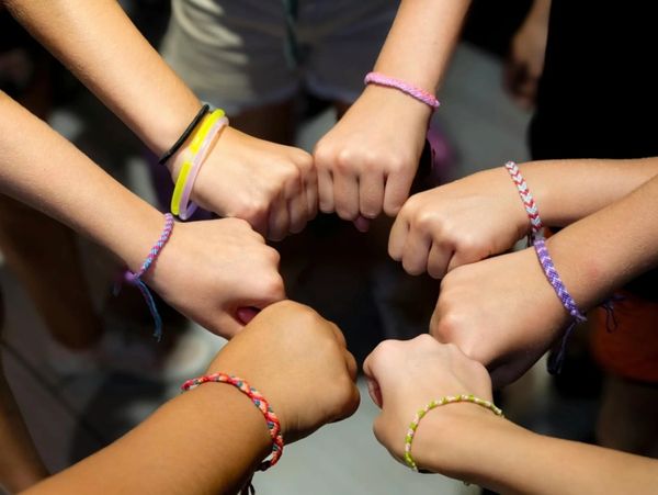 A group of six hands forming a fist bump circle, each wearing colorful friendship bracelets.