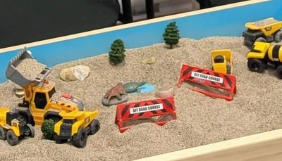 Toy construction vehicles and roadblocks in a sandbox with small trees.