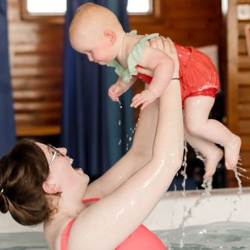A woman joyfully lifts a baby out of the water in a pool.
