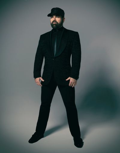 Man in an elegant black suit and cap poses confidently against a grey backdrop.
