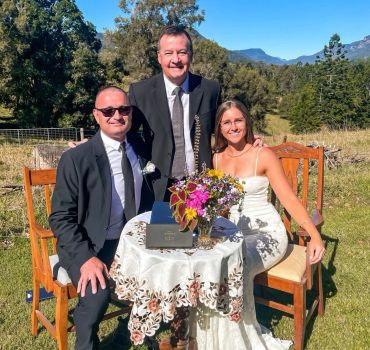 Three people in formal attire around a small table outdoors.