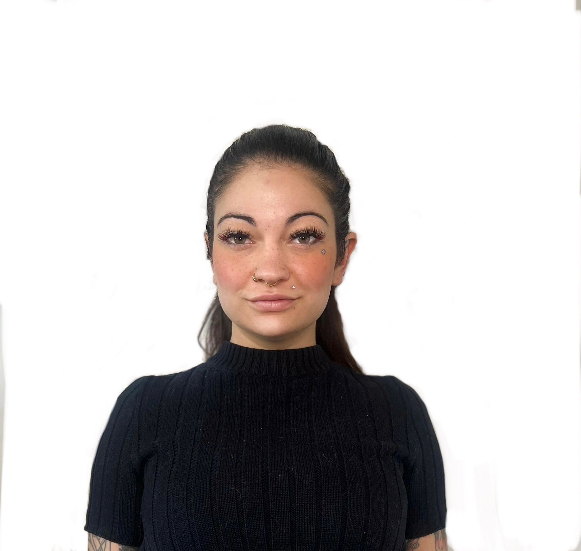 A confident woman with facial piercings and a black ribbed top against a white background.
