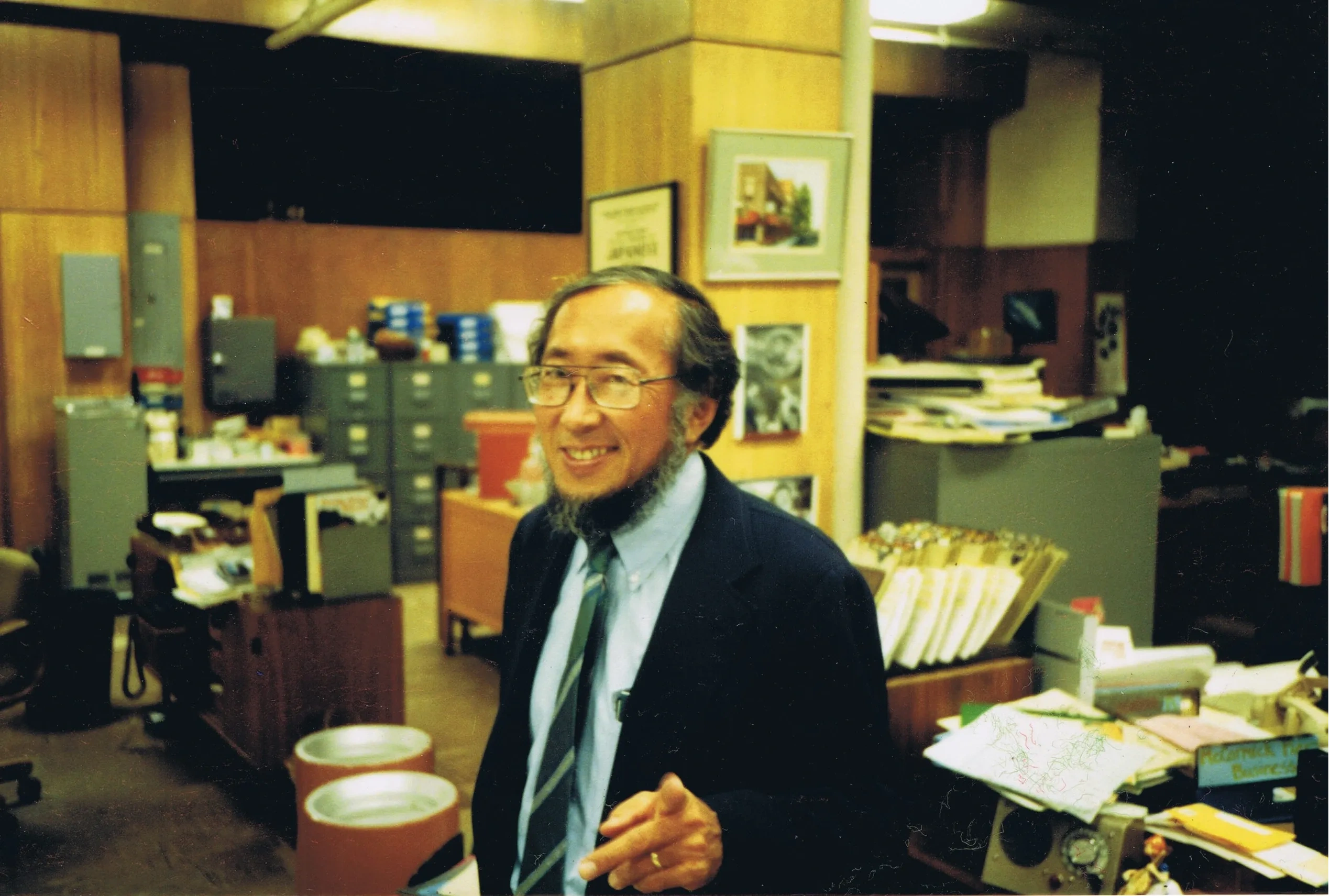 Older man in offices with lots of papers and desks pointing at the camera with a smile