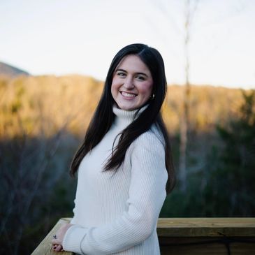 Young woman in a white sweater smiling outdoors with nature background.