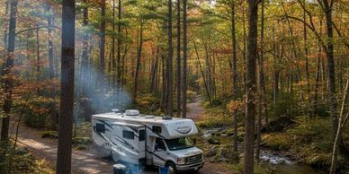 RV camping by a peaceful forest stream with a campfire and chairs.