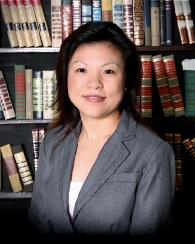 A woman with dark hair wearing a dark gray blazer, standing in front of a book case.