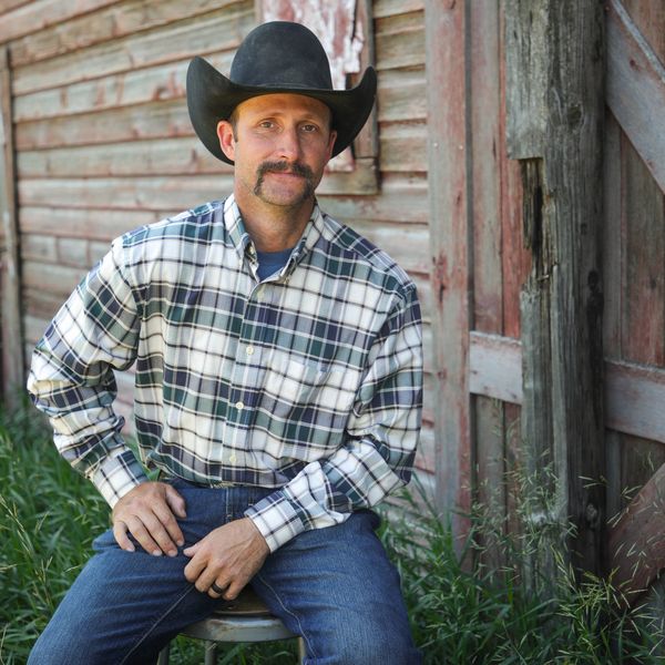 Man in a cowboy hat sitting outside a rustic wooden building.