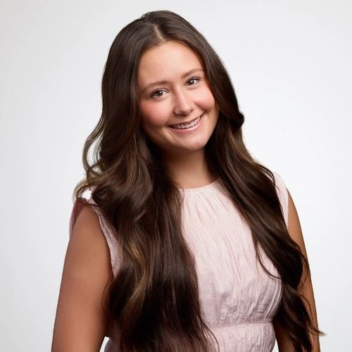 Smiling young woman with long wavy brown hair in a light pink top.