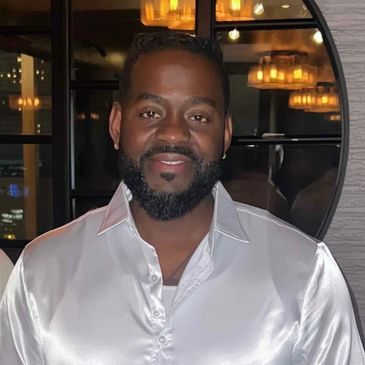 Man smiling in a white satin shirt at a dimly lit venue.