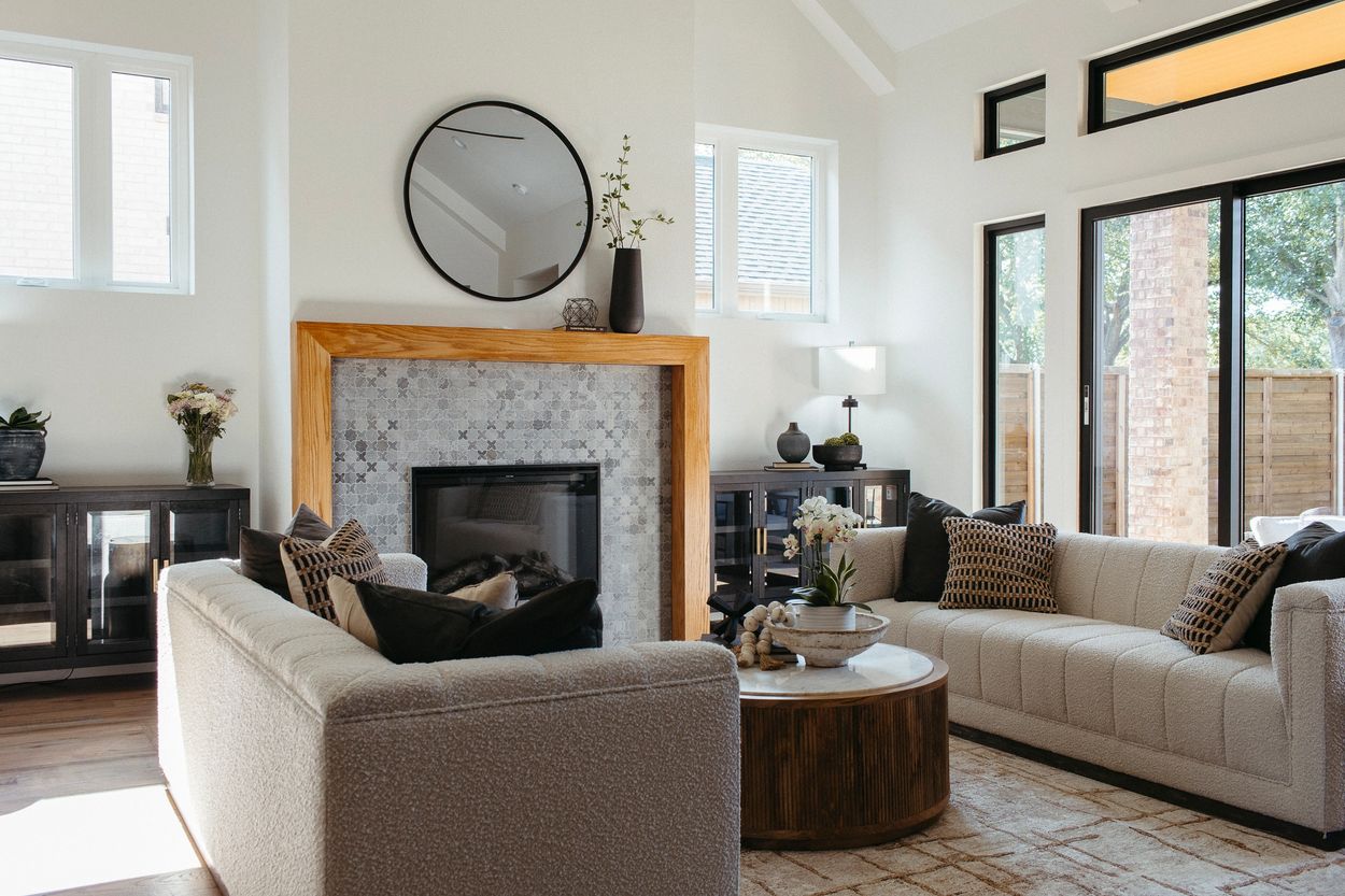 Grapevine family room with stone fireplace, warm wood mantel, round mirror, neutral sofas, and black-framed windows.