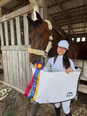 Young equestrian proudly holding ribbons and a saddle pad beside her horse in a stable.