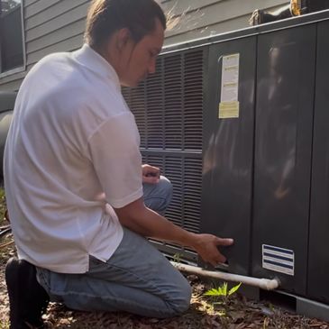 A technician inspects an HVAC unit outdoors, kneeling on the ground.