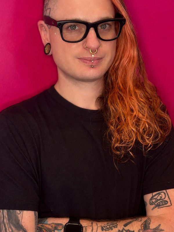 Person with long orange hair, black glasses, and facial piercings against a pink background.