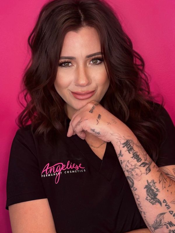 A woman with dark hair and tattoos poses against a pink background.