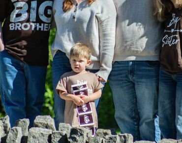 A young boy stands with family members wearing sibling-themed shirts outdoors.