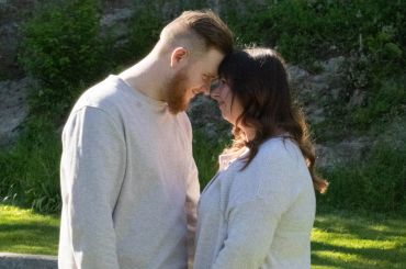 A couple in light sweaters touching foreheads affectionately outdoors.