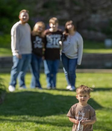 Young boy stands in front wearing a 'big brother' shirt with family blurred in the background.