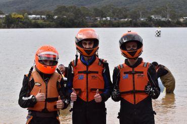 Three people in orange helmets and life jackets giving thumbs up by the water.