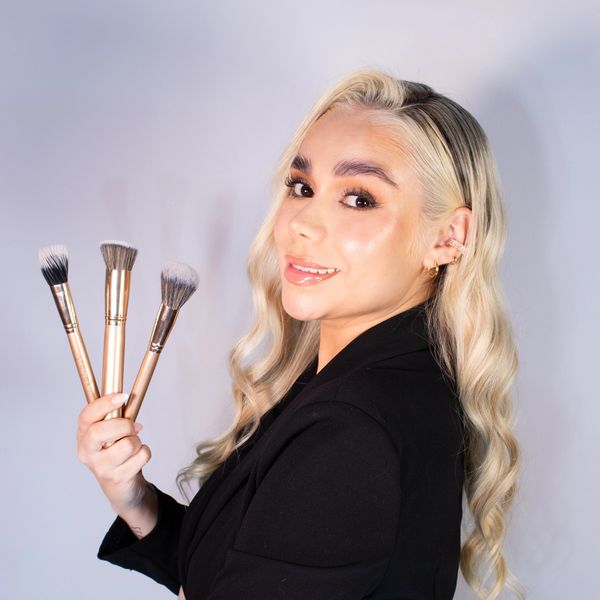 Woman with long blonde hair holding three makeup brushes, smiling.