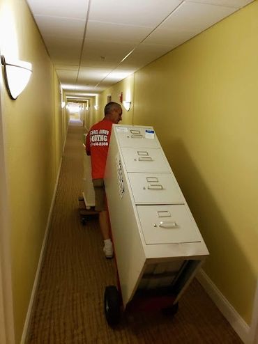 Mover transporting a large filing cabinet down a narrow hallway.