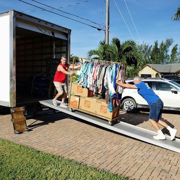 Two men loading a rack of clothes and boxes into a truck using a ramp.