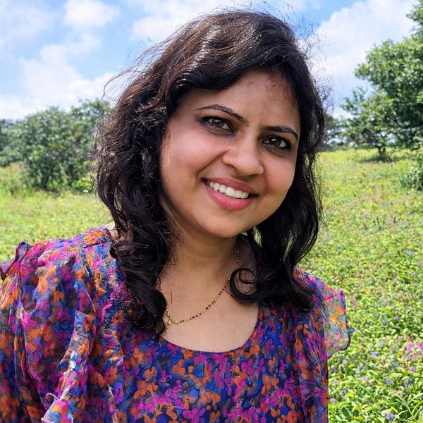 A woman smiling outdoors in a colorful floral dress with greenery and blue sky behind her.
