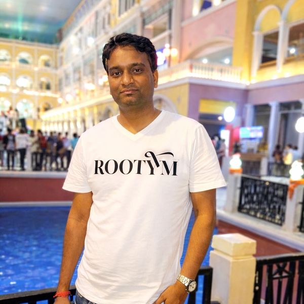 Man in a white ROOTYM t-shirt posing indoors by a blue water feature.