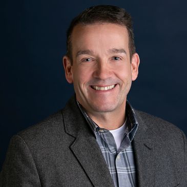 Smiling man in a gray blazer and plaid shirt with a dark blue background.