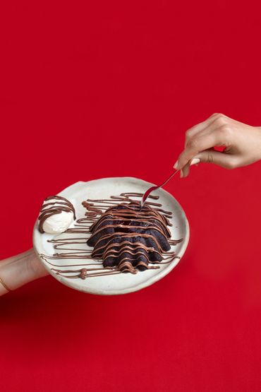 chocolate volcano cake on a plate with hands reaching out to eat it. background is bright red
