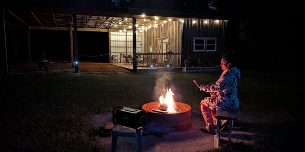 Person warming hands by a fire pit at night near a lit cabin.