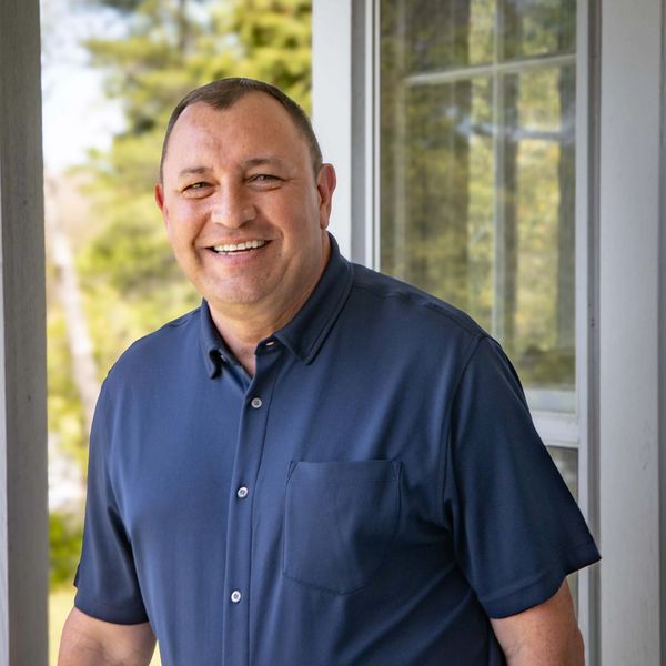 Smiling man in a blue shirt standing outdoors near a window.
