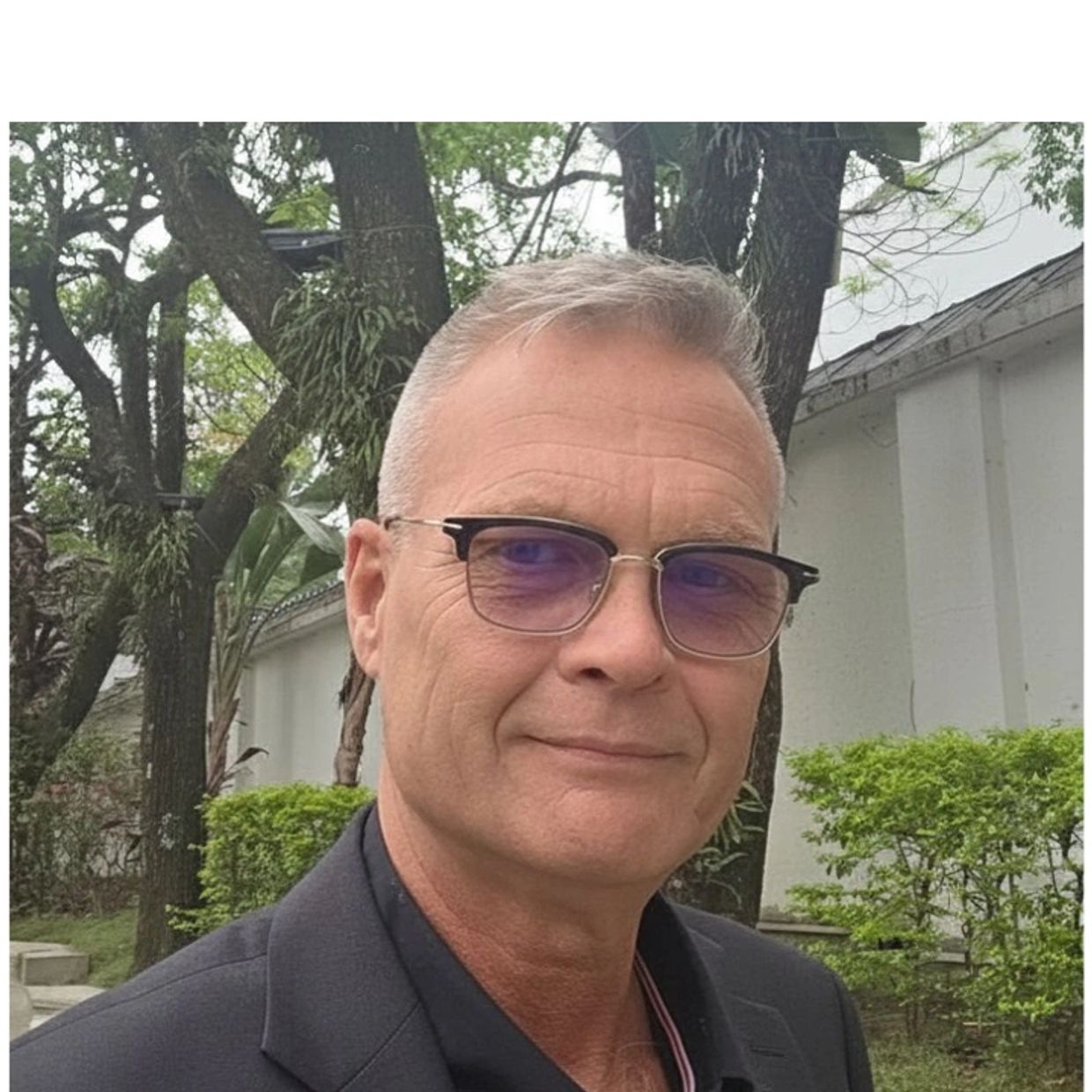 Smiling man wearing glasses and a dark blazer outdoors.