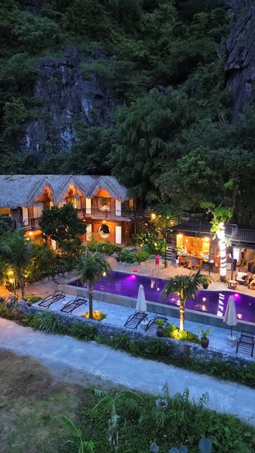 A cozy resort with a lit pool nestled against rocky cliffs and lush greenery at dusk.